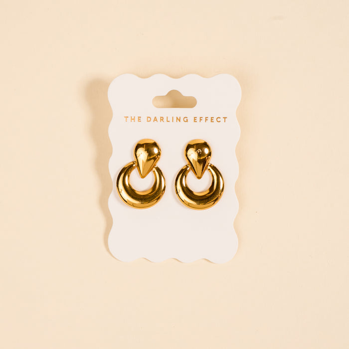 The Heirloom Standout Earrings by The Darling Effect, featuring a teardrop-shaped top and high-shine finish, are displayed on a white scalloped card with the brand name against a beige background.