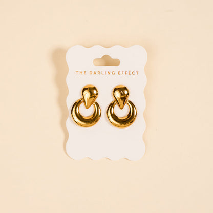 The Heirloom Standout Earrings by The Darling Effect, featuring a teardrop-shaped top and high-shine finish, are displayed on a white scalloped card with the brand name against a beige background.