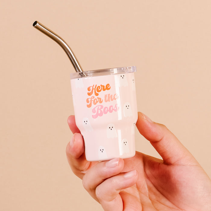 A hand holds The Darling Effect's "Here for the Boos" 2 oz Tiny Tumbler, a pink cup with ghost designs and orange and white lettering, insulated for festive sips—perfect as a Halloween-themed shot glass.