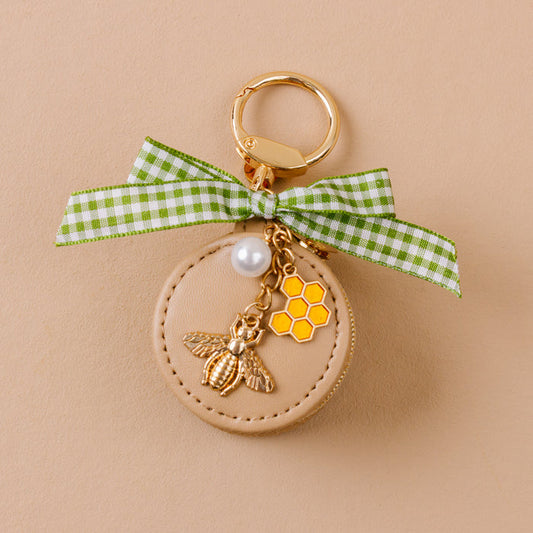 The Darling Effect’s Hey Honey Travel Jewelry Case Bag Charm features a beige round keychain with a gold bee, honeycomb charm, pearl, and green gingham ribbon on a gold keyring—styled against a light tan background.