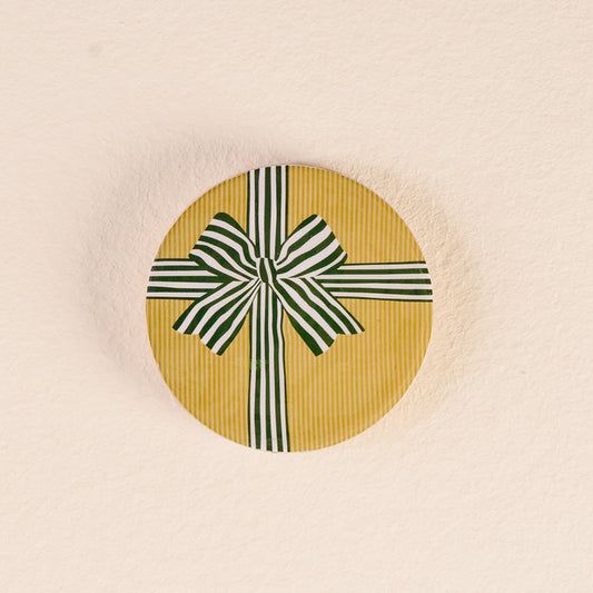 The Darling Effect’s Holiday Gift Bow Green Wood Enamel Coaster comes in a round yellow box with subtle stripes and a bold green-and-white bow, perfect for gifting, set against a textured light beige background.