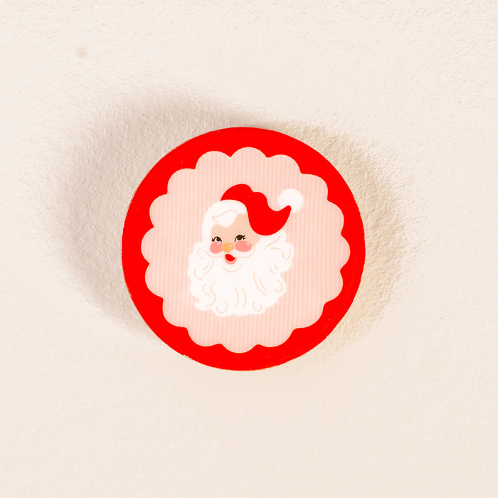 Add festive charm to your decor with The Darling Effect's Holly Jolly Santa Wood Enamel Coaster, featuring a round design with a red border and cartoon Santa Claus face—perfect for any holiday coaster set.