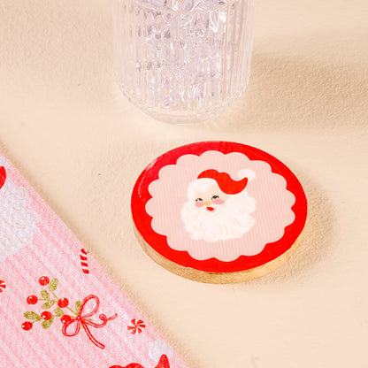 A round cookie with a Santa face on red icing rests on a beige surface beside The Darling Effect's Holly Jolly Santa Wood Enamel Coaster, a clear glass, and a pink holiday cloth with candy and bows.