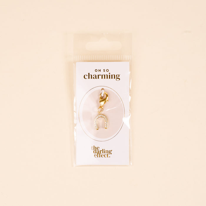 The-darling-effect Horseshoe Jewelry Charm Gold features small crystals, attached to an "OH SO charming" card—perfect for your charm bar. Unique jewelry packaged in a clear plastic bag against a beige background.