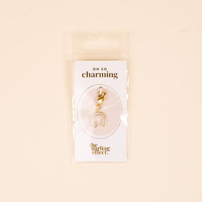 The-darling-effect Horseshoe Jewelry Charm Gold features small crystals, attached to an "OH SO charming" card—perfect for your charm bar. Unique jewelry packaged in a clear plastic bag against a beige background.