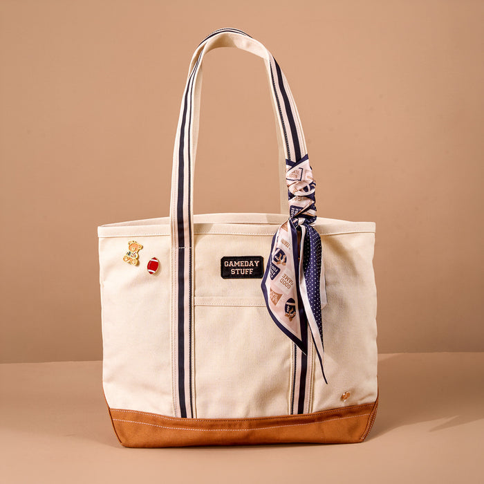 A cream Huddle Up The Darling Canvas Tote Bag by The Darling Effect, featuring a brown bottom, navy stripes, a "GAMEDAY STUFF" patch, two pins, and a knotted patterned scarf on the handle, is displayed against a plain beige background. Adorned with embroidered patches and designed with a roomy interior, this tote seamlessly combines style and practicality.