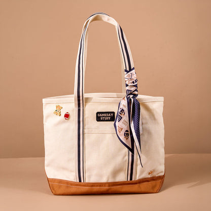 A cream Huddle Up The Darling Canvas Tote Bag by The Darling Effect, featuring a brown bottom, navy stripes, a "GAMEDAY STUFF" patch, two pins, and a knotted patterned scarf on the handle, is displayed against a plain beige background. Adorned with embroidered patches and designed with a roomy interior, this tote seamlessly combines style and practicality.