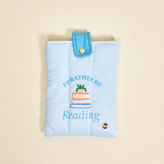 The Darling Effect's "I'd Rather Be Reading Book Cover" in light blue polyester features a quilted design, snap closure, vibrant stacked books with a plant, and protects your books in cute style.