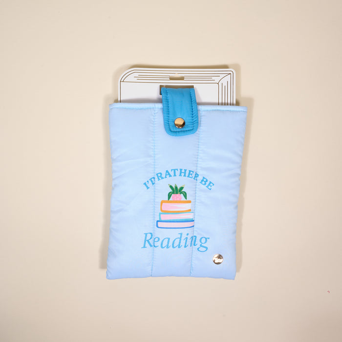 The Darling Effect's "I'd Rather Be Reading" Book Cover is a light blue, puffy polyester pouch with colorful embroidered books—perfect for protecting your reads. Shown against a cream background.