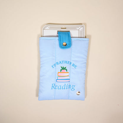 The Darling Effect's "I'd Rather Be Reading" Book Cover is a light blue, puffy polyester pouch with colorful embroidered books—perfect for protecting your reads. Shown against a cream background.