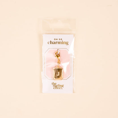 The-darling-effect J Jewelry Charm Initial Letter Gold features a gold-plated brass "J" with a glitter border, clasp, and is displayed on a branded card in clear packaging—perfect personalized jewelry on a cute pink-and-white background.