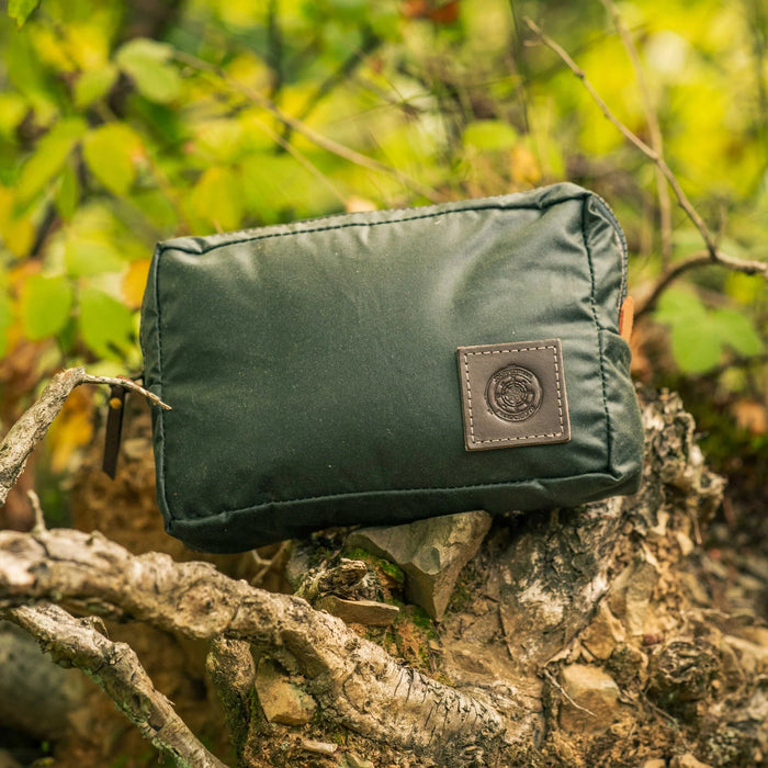 Forager Provisions LTD green waxed canvas expedition pouch with a leather logo patch, resting on a mossy forest log.