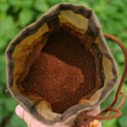 Hand holding Journeyman Craft Khaki Brown Waxed Canvas Coffee Pouch full of ground coffee, greenery in blurred background.
