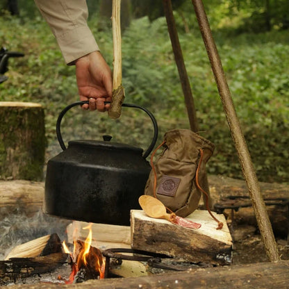 Camper brews coffee over campfire with black kettle, khaki coffee pouch, and spoon by Journeyman Craft resting on a log.