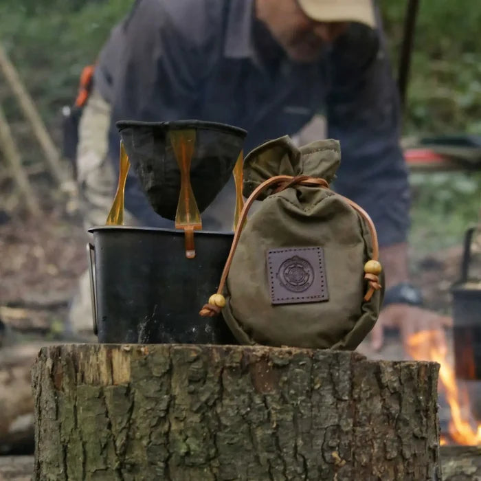 Khaki green Journeyman Craft waxed canvas coffee pouch rests on a stump by a glowing campfire in the great outdoors.