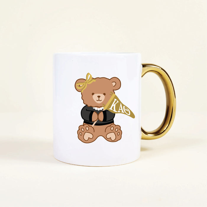 The Darling Effect’s Kappa Alpha Theta 11 oz Cozy Cute Ceramic Mug features a charming teddy bear in a black top, pearl necklace, yellow bow, and gold handle, holding a yellow KAO flag—perfect for sorority members.