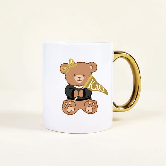 The Darling Effect’s Kappa Alpha Theta 11 oz Cozy Cute Ceramic Mug features a charming teddy bear in a black top, pearl necklace, yellow bow, and gold handle, holding a yellow KAO flag—perfect for sorority members.