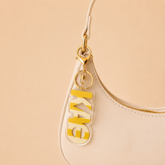 A beige handbag with a gold zipper, featuring a gold keyring holding The Darling Effect’s Kappa Alpha Theta Stripes Acrylic Keychain—a clear tag with bold yellow KAE letters—makes the perfect sorority gift for bid day.