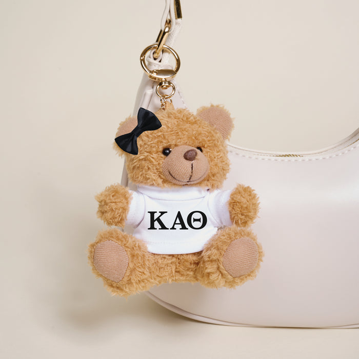 The Darling Effect's Kappa Alpha Theta Teddy Bear Bag Charm—a small brown bear with a black bow and KAΘ shirt—hangs on a white handbag, making it the perfect Bid Day accessory.