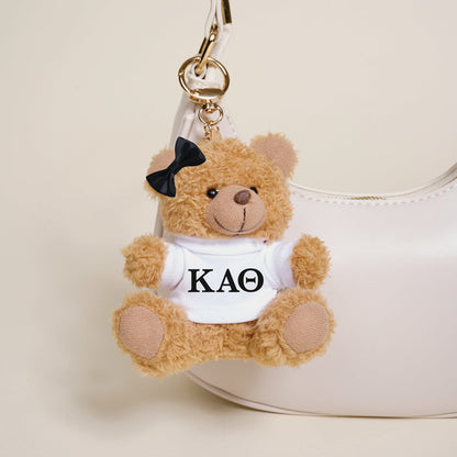 The Darling Effect's Kappa Alpha Theta Teddy Bear Bag Charm—a small brown bear with a black bow and KAΘ shirt—hangs on a white handbag, making it the perfect Bid Day accessory.