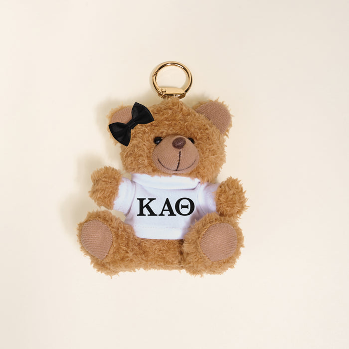 The Darling Effect’s Kappa Alpha Theta Teddy Bear Bag Charm features a plush brown bear with a black bow on its left ear, wearing a white shirt with “KAΘ” in black—an adorable Bid Day accessory, shown on a cream background.