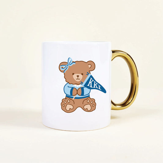 The Darling Effect’s Kappa Kappa Gamma 11 oz Cozy Cute Ceramic Mug features a white base, gold handle, and a brown teddy bear in blue clothes with Greek letters ΚΚΓ—perfect for any Kappa Kappa Gamma member.