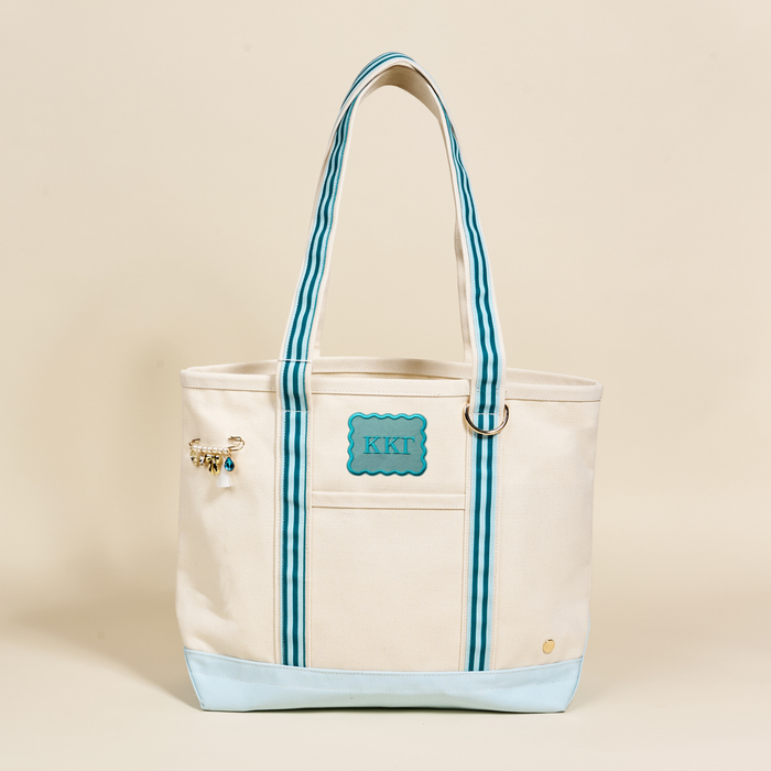 The Darling Effect’s Kappa Kappa Gamma Canvas Tote Bag features a cream canvas body, blue striped handles, a light blue base, a blue KKT patch with sorority letters, and a gold charm on one handle—all set against a plain background.