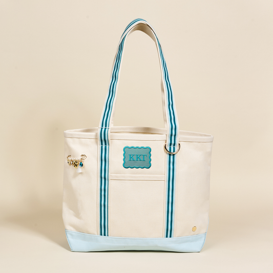 The Darling Effect’s Kappa Kappa Gamma Canvas Tote Bag features a cream canvas body, blue striped handles, a light blue base, a blue KKT patch with sorority letters, and a gold charm on one handle—all set against a plain background.