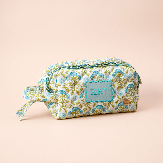 The Darling Effect’s Kappa Kappa Gamma Essentials Quilted Cosmetic Pouch features a floral design, zipper, side handle, and embroidered KKG on a light blue label—ideal for storing sorority must-haves on a pale peach background.