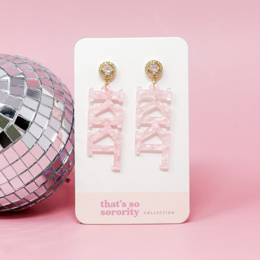 A pair of Kappa Kappa Gamma Letter Pink Acrylic Earrings by The Darling Effect hang from a white card labeled "that's so sorority collection" with a pink background—ideal for sisterhood events or bid-day gifts. A disco ball peeks in on the left.