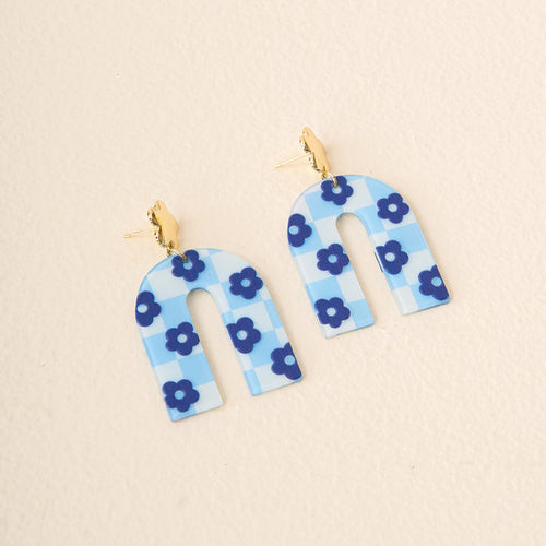 Light Blue Game Day Acrylic Earrings by The Darling Effect feature arched acrylic with a blue and white checkered pattern, blue flower accents, and gold stud posts. Perfect for showing school spirit. Displayed on a light beige surface.