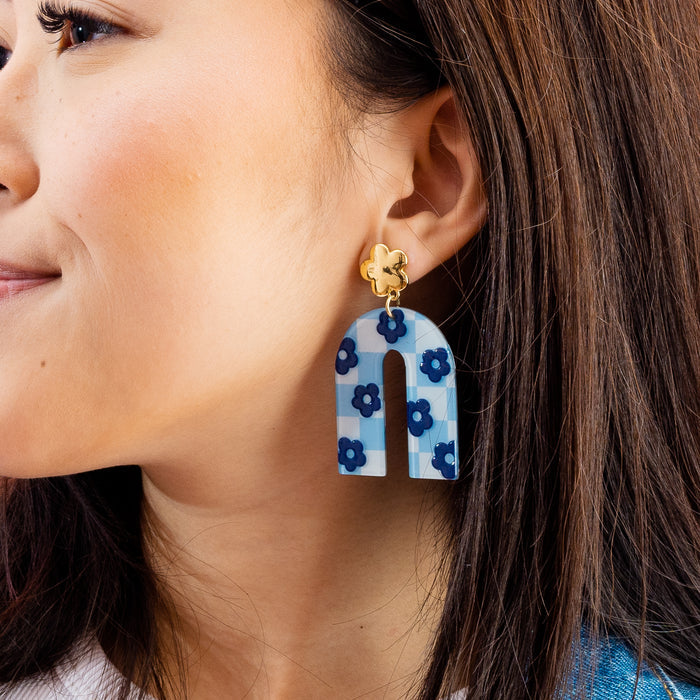 A close-up of a woman wearing The Darling Effect's Light Blue Game Day Acrylic Earrings, featuring a large blue-and-white checkered U-shaped design with dark blue flowers and a gold flower-shaped stud—perfect for showcasing school spirit.