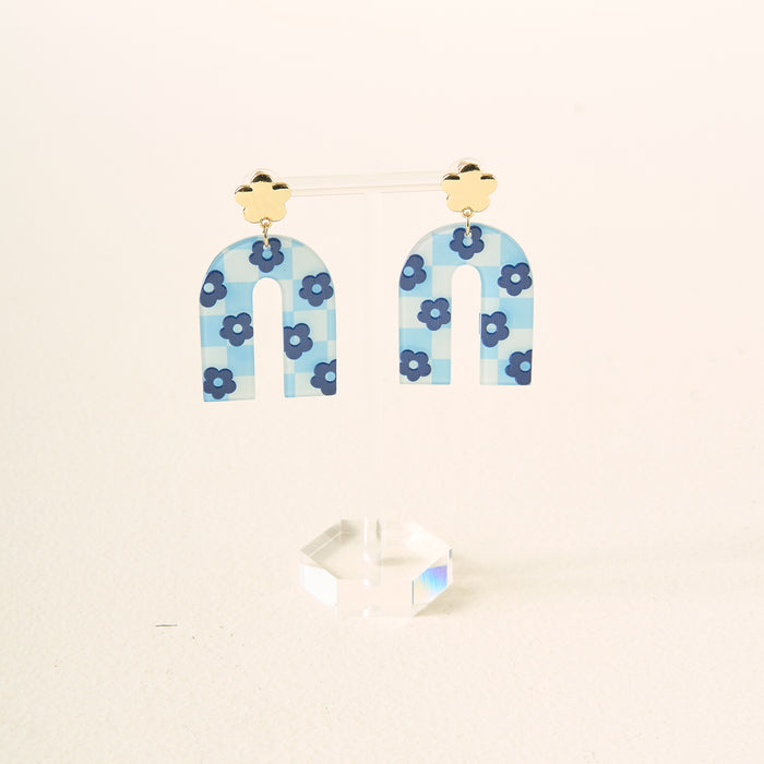 The Darling Effect's Light Blue Game Day Acrylic Earrings feature arch shapes in blue tones with a checkered pattern, small blue flowers, and gold-colored flower studs. Displayed on a clear stand with a white background—perfect for school spirit or game day.