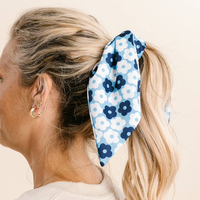 A woman with blonde hair in a ponytail wears The Darling Effect’s Light Blue Game Day Hair Scarf with Scrunchie, paired with gold hoop earrings, as she’s shown from the side against a neutral background.
