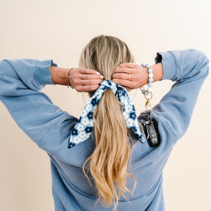 A woman with long blonde hair, tied back with The Darling Effect’s Light Blue Game Day Hair Scarf with Scrunchie, faces away from the camera. She wears a blue sweatshirt and holds car keys on a beaded keychain—perfect for showing team spirit.