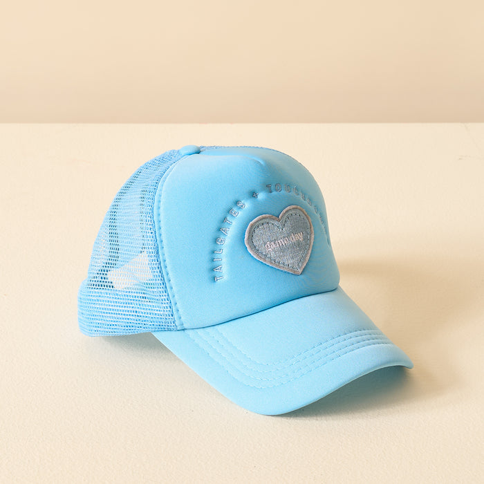 The Darling Effect’s Light Blue Game Day Trucker Hat features mesh panels, a heart patch, and embroidered text on the front—displayed on a cream surface with a beige backdrop. Perfect for showing your team spirit!.