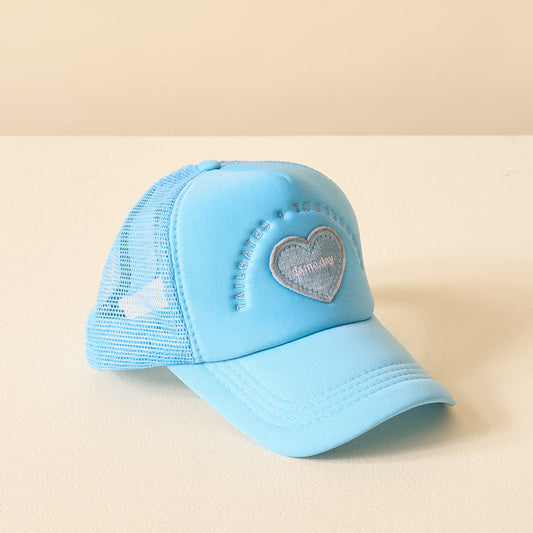 The Darling Effect’s Light Blue Game Day Trucker Hat features mesh panels, a heart patch, and embroidered text on the front—displayed on a cream surface with a beige backdrop. Perfect for showing your team spirit!.