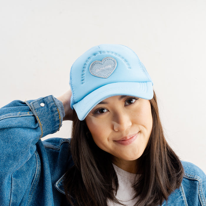 A woman with long dark hair wears The Darling Effect’s Light Blue Game Day Trucker Hat, a white top, and a blue denim jacket. She smiles softly, touching the back of her head while posing against a plain white background.