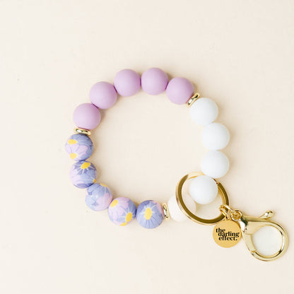 The Lilac Bloom Hands-Free Keychain Wristlet by the-darling-effect features lavender and white silicone beads, floral accents, and a gold clasp for stylish, hands-free carrying.