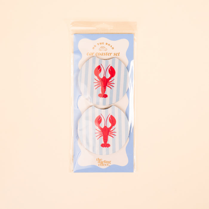The Darling Effect Lobster On the Road Car Coaster Set includes two round absorbent coasters with blue stripes and red lobster designs, packaged in a clear bag with a light blue card—perfect for your car cup holder collection.