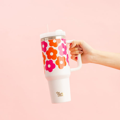 A hand holds The Darling Effect's Lovebug Floral 40 oz Take Me Everywhere Tumbler, featuring orange and pink retro flowers, a handle, straw, and gold "the drift effect" text near the bottom, set against a light pink background.