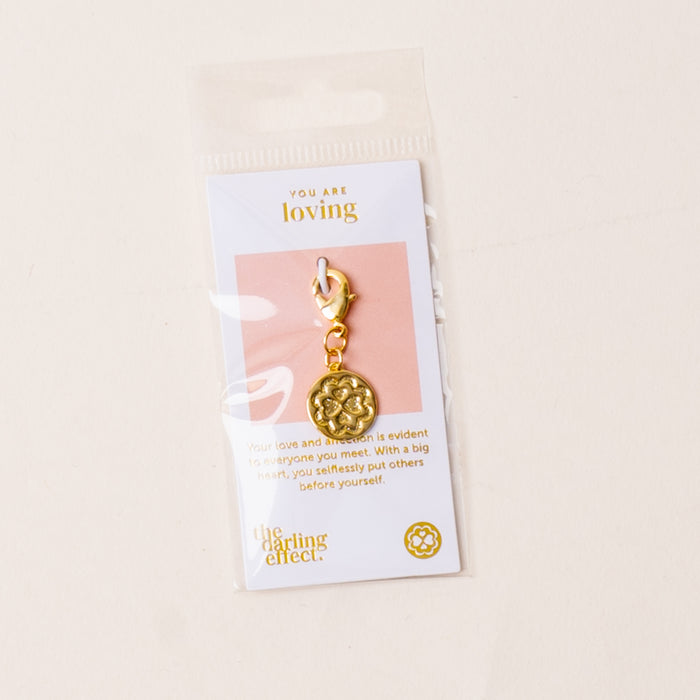 The-darling-effect’s Loving Jewelry Trait Charm Gold features a floral design on a card stating “You are loving.” Packaged in a clear plastic bag, it celebrates strength and selfless love with its heartfelt message.