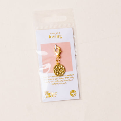 The-darling-effect’s Loving Jewelry Trait Charm Gold features a floral design on a card stating “You are loving.” Packaged in a clear plastic bag, it celebrates strength and selfless love with its heartfelt message.