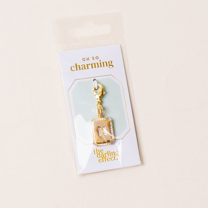 The-darling-effect’s M Jewelry Charm Initial Letter Gold features a gold-plated brass “M” with rhinestones, displayed in clear packaging labeled OH SO charming. Ideal for personalized jewelry or as an initial letter charm. Background is light and neutral.
