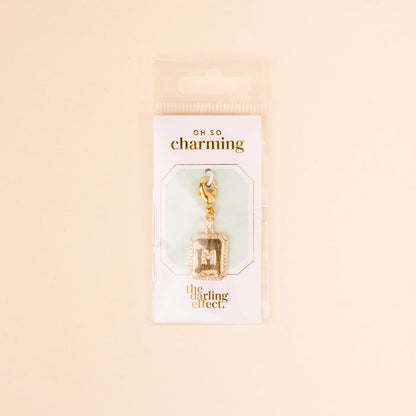 The M Jewelry Charm Initial Letter Gold by the-darling-effect features a gold-plated charm attached to a lobster clasp, displayed on white Oh So Charming packaging—an ideal gift for personalized jewelry lovers.
