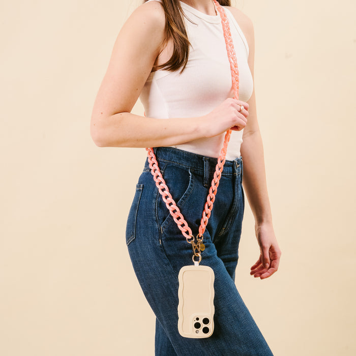 A woman in a sleeveless white top and blue jeans holds her phone in a beige case with the-darling-effect’s Marble Bubblegum Hold the Phone Crossbody Chain, worn stylishly across her body against a plain beige background.