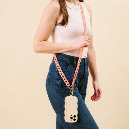 A woman in a sleeveless white top and blue jeans holds her phone in a beige case with the-darling-effect’s Marble Bubblegum Hold the Phone Crossbody Chain, worn stylishly across her body against a plain beige background.