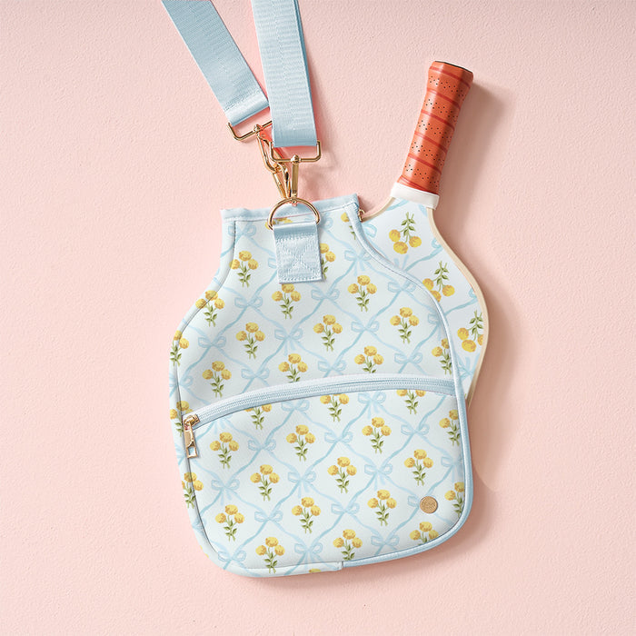 The Marigold Lane Blue Pickleball Paddle Case by the-darling-effect, featuring a yellow floral pattern and gold zipper, holds a paddle with an orange grip and hangs from a matching blue strap against a pale pink background.