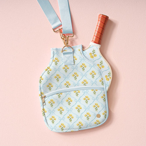 The Marigold Lane Blue Pickleball Paddle Case by the-darling-effect, featuring a yellow floral pattern and gold zipper, holds a paddle with an orange grip and hangs from a matching blue strap against a pale pink background.