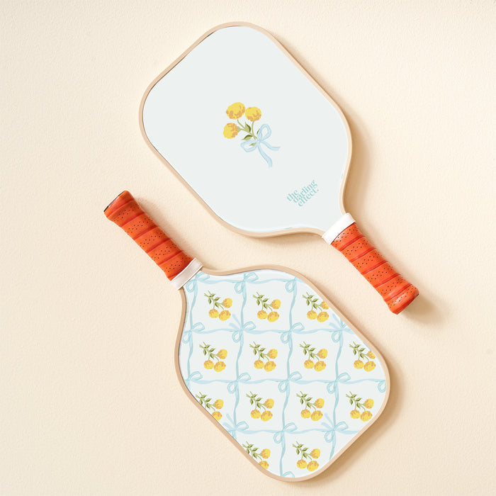 The-darling-effect's Marigold Lane Blue Pickleball Paddle features orange handles and lively designs: one with a yellow flower bouquet tied by a blue ribbon, the other with playful yellow flowers and blue ribbon patterns.